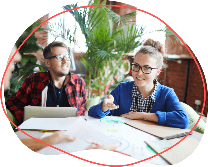 Dvaja ľudia s okuliarmi diskutujú pri stole s notebookom o papieroch, obklopení bujnými rastlinami a zvažujú vplyv poukážky na ich projekt.