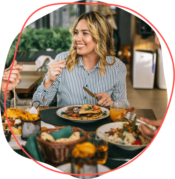 Žena v pruhovanej košeli sa pri stolovaní s priateľmi v reštaurácii usmieva, vychutnáva si jedlo a diskutuje o tom, ako zvládnuť stravné na nadchádzajúcu cestu.