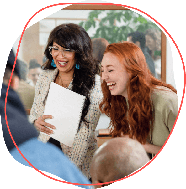 Dve ženy, ktoré sa spolu smejú, jedna drží notebook a zdieľajú výhody svojich zážitkov v prostredí živej skupiny.