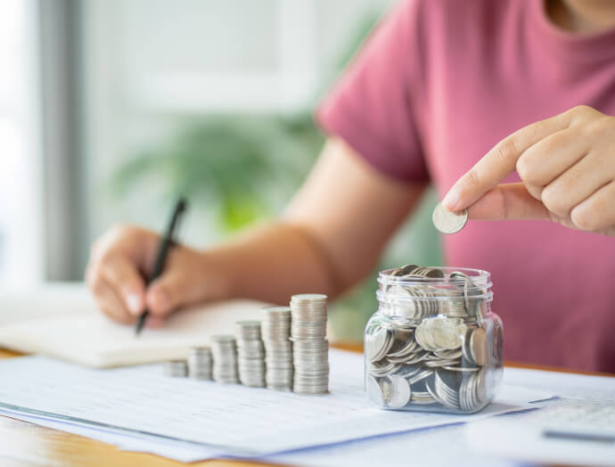 Osoba v ružovej košeli s precíznosťou ukladá mince, ukladá ich do téglika, zatiaľ čo si zapisuje poznámky ako správny Finmajster pri svojom drevenom stole.